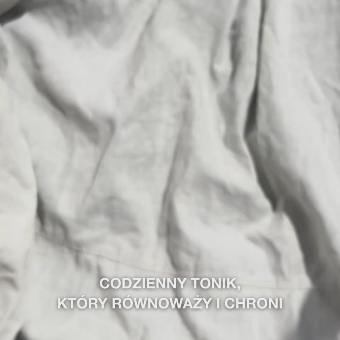 Zbliżenie ręki, która nakrapia kilka kropli przezroczystego, wodnistego toniku Face Tonic z olejkiem z drzewa herbacianego na wacik, a następnie delikatnie przeciera nim oczyszczoną skórę twarzy: lekki, orzeźwiający płyn łagodzi, głęboko oczyszcza, nawilża i pozostawia skórę odnowioną i gotową na krem.